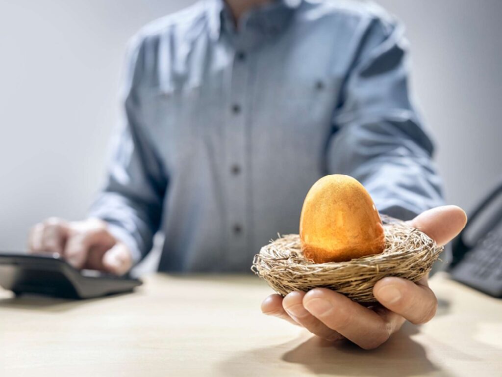 Businessman holds nest with golden egg inside whilst typing on a calculator at a desk with a phone next to him