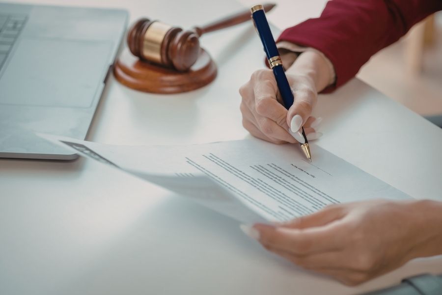 A person signing a legal document with a pen. The document is placed on a table, and a gavel is positioned nearby, suggesting a formal or judicial setting.